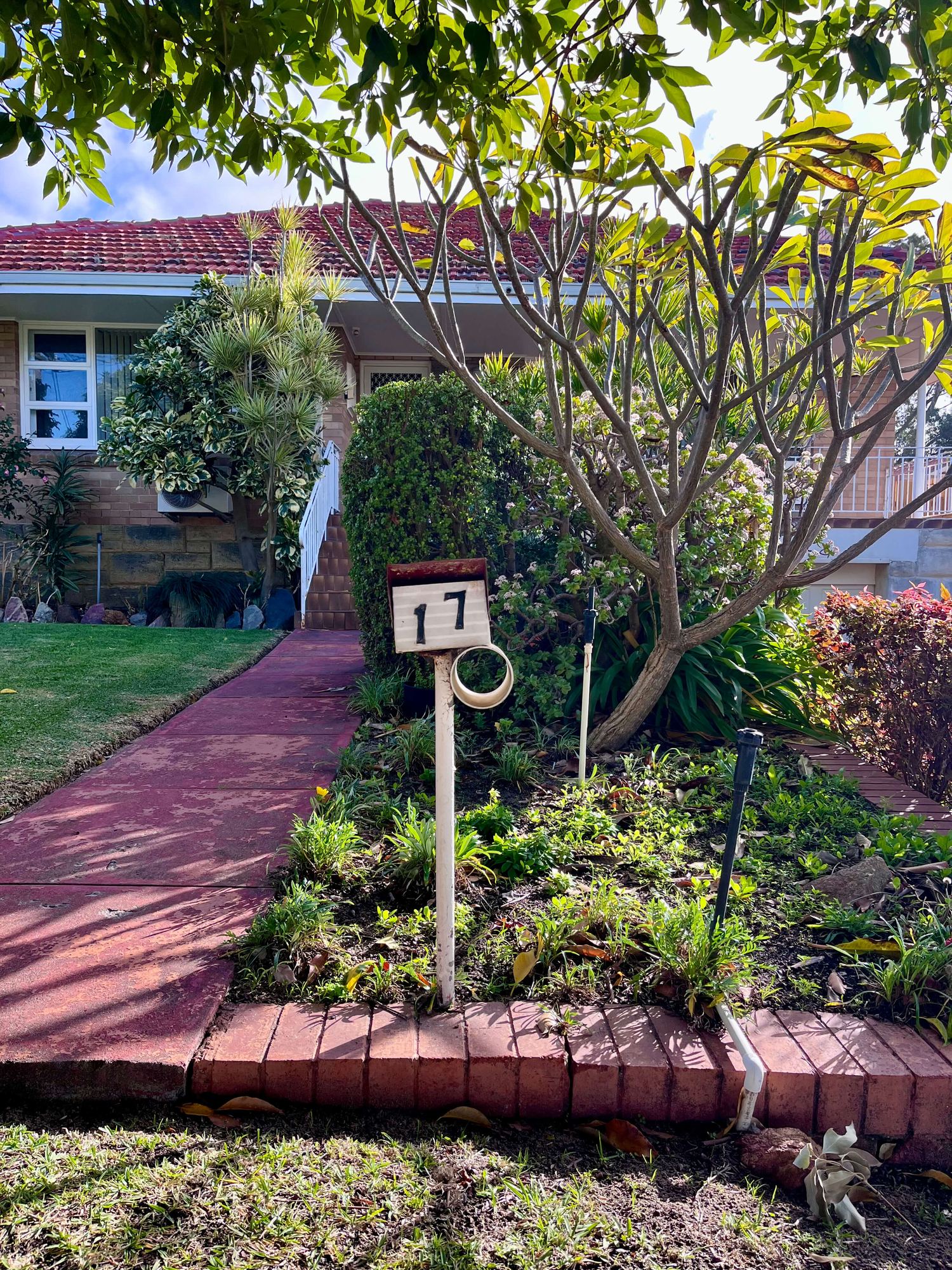 A rusted circular letterbox marked 17, ringed by a garden of shrubs and a frangipani tree