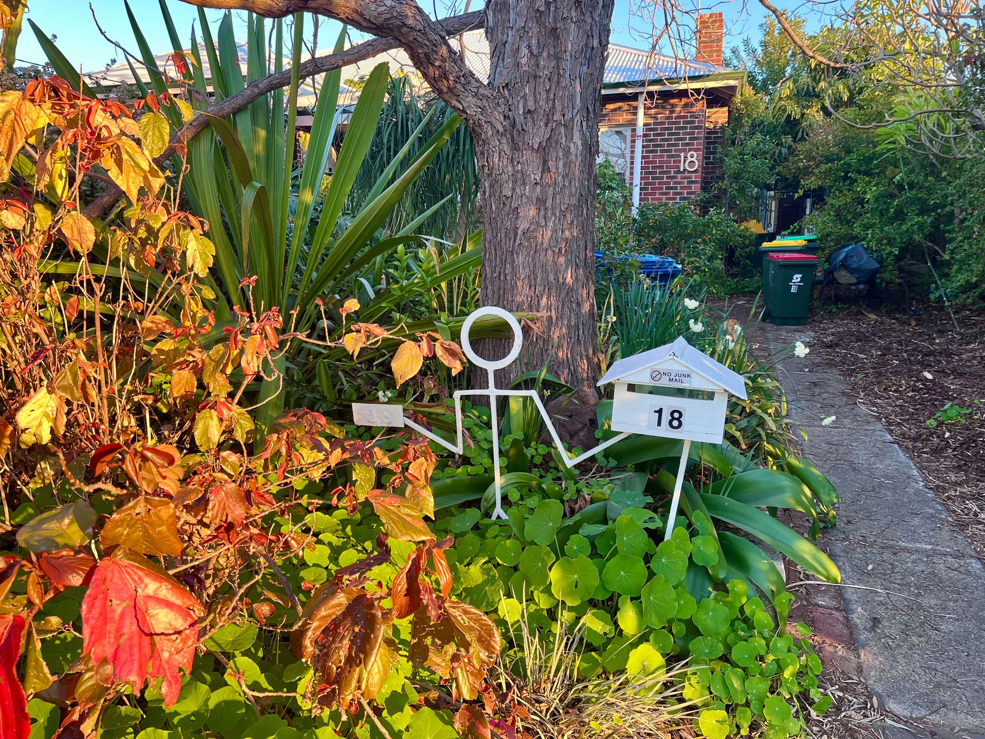 A whimsical stick-figure sculpture holding up a small letterbox marked 18, next to a tree trunk