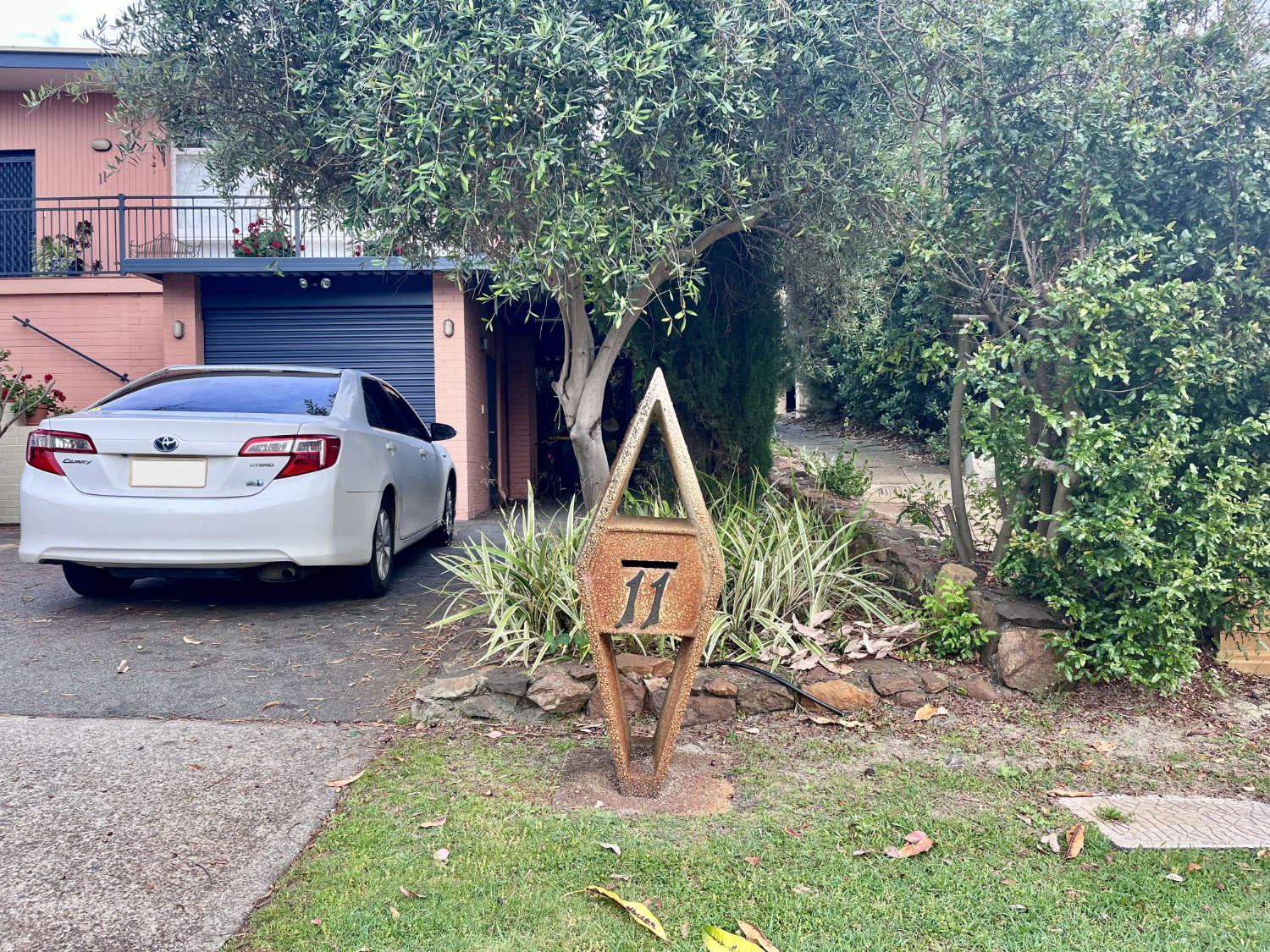 A rusted, diamond-shaped letterbox marked 11, standing beside an olive tree near the driveway